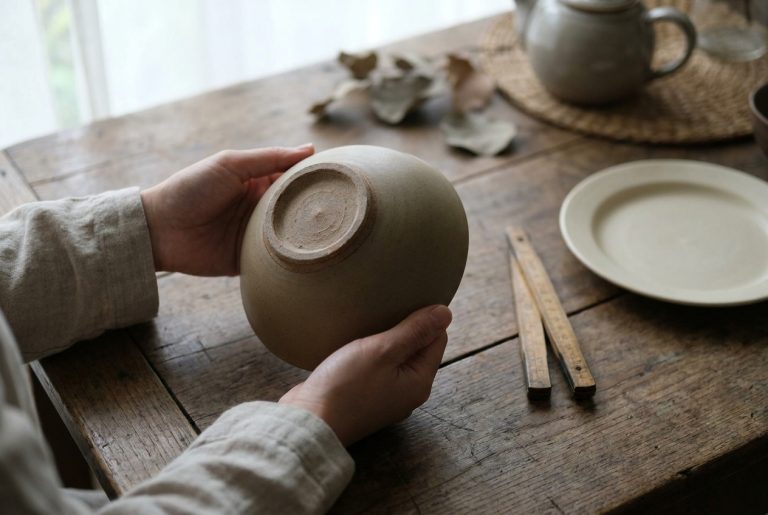Hands turning a plain Japanese ceramic bowl to examine its unglazed foot ring, with a folding ruler on a wooden table
