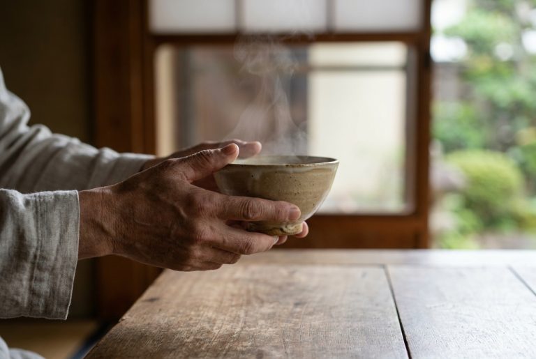 Hands lifting a small ceramic rice bowl from a wooden table, fingers supporting the foot ring