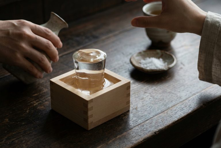 A plain hinoki masu box on a dark counter with a glass tumbler set inside it, sake brimming over the rim and pooling in the box, a small dish of salt behind